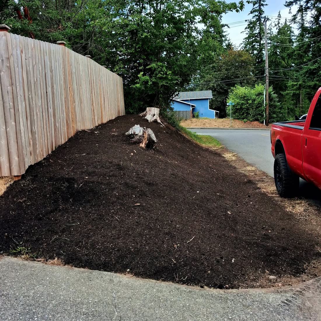 Fresh dark mulch spread over a slope beside a new wooden fence, a red pickup at the curb.